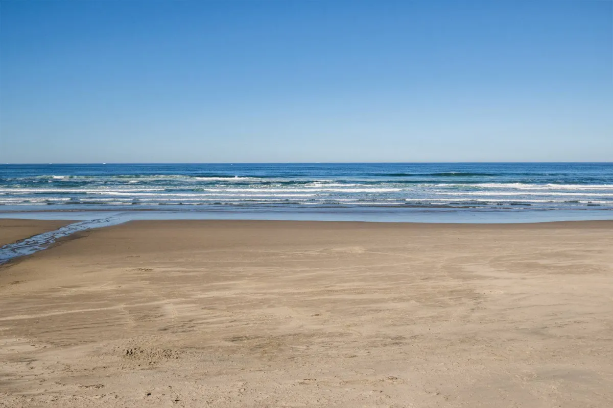 A scenic view of a sandy beach with gentle ocean waves rolling in and a clear blue sky above. The tranquil setting is ideal for sun-filled days, sandcastle building, and sunset strolls, inviting families to enjoy the natural beauty and carefree activities the Oregon Coast offers.