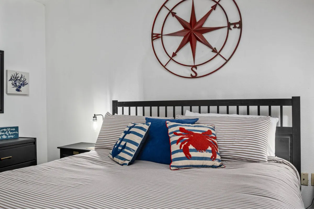 A loft bedroom with striped bedding, a decorative red crab pillow, and a compass motif on the wall. Ample natural light streams in, creating an airy and inviting atmosphere ideal for unwinding.