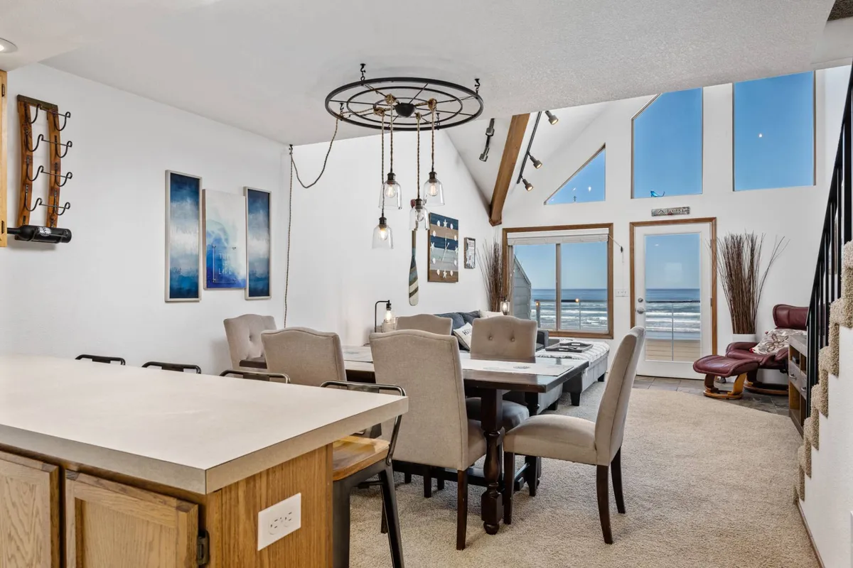 A warmly lit dining area features a central table perfect for family dinners or social gatherings, with large windows in the background offering an inviting ocean view. The chandelier and decor lend a boutique inn feel to the space.