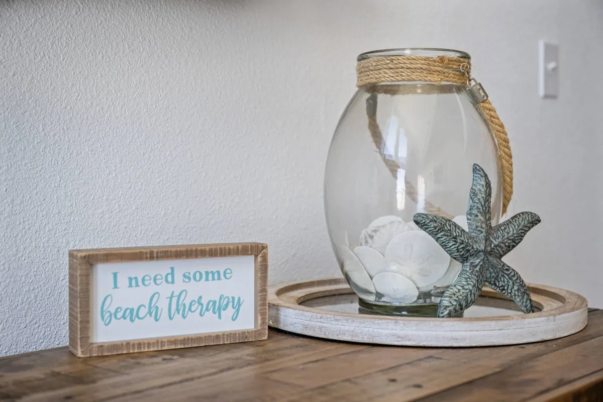 This image features a tranquil nook adorned with a glass jar of seashells, a starfish decoration, and a wooden sign, all set against a softly lit background. The decor captures the essence of the coast and invites moments of quiet reflection and inspiration, with an unmistakable sense of beachside calm.