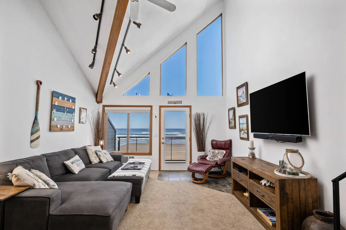 The image showcases a bright, inviting living area with a cozy sectional sofa facing expansive windows that reveal a sweeping ocean view. A wooden entertainment center with a mounted TV creates a comfortable, homey atmosphere, while the natural light and panoramic seascape set a tranquil and uplifting mood. The setup is perfect for relaxing, socializing, or simply unwinding to the soothing sounds of the surf.
