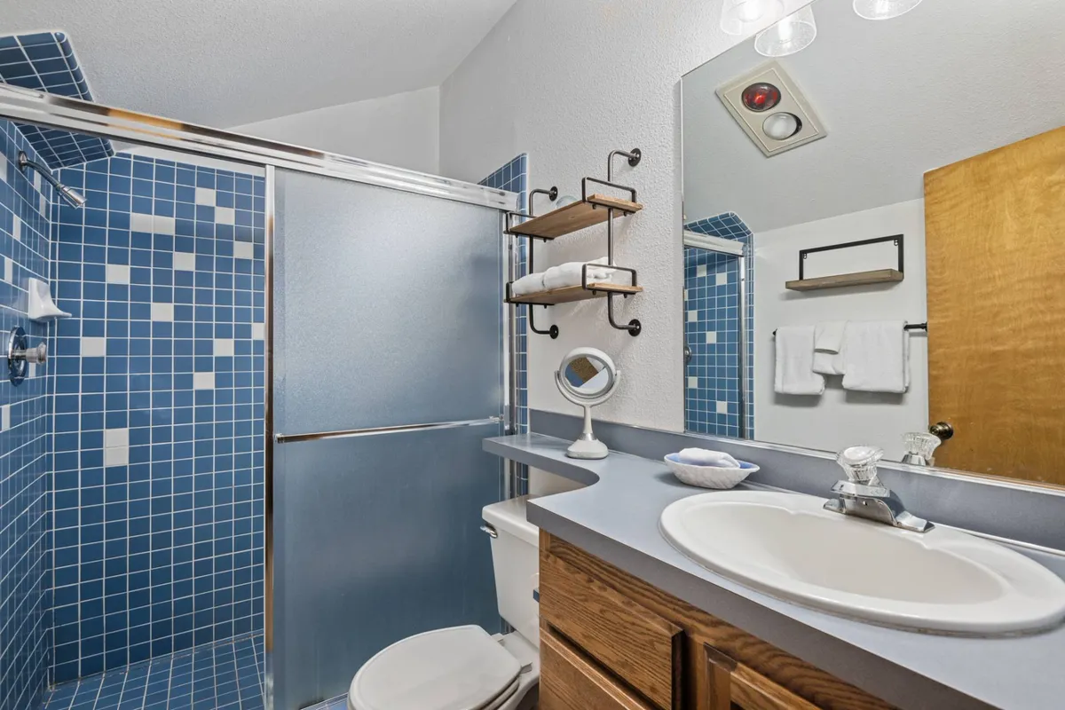 A modern en-suite bathroom with a blue tiled walk-in shower, a sleek vanity with sink, large mirror, and wooden shelves for storage.