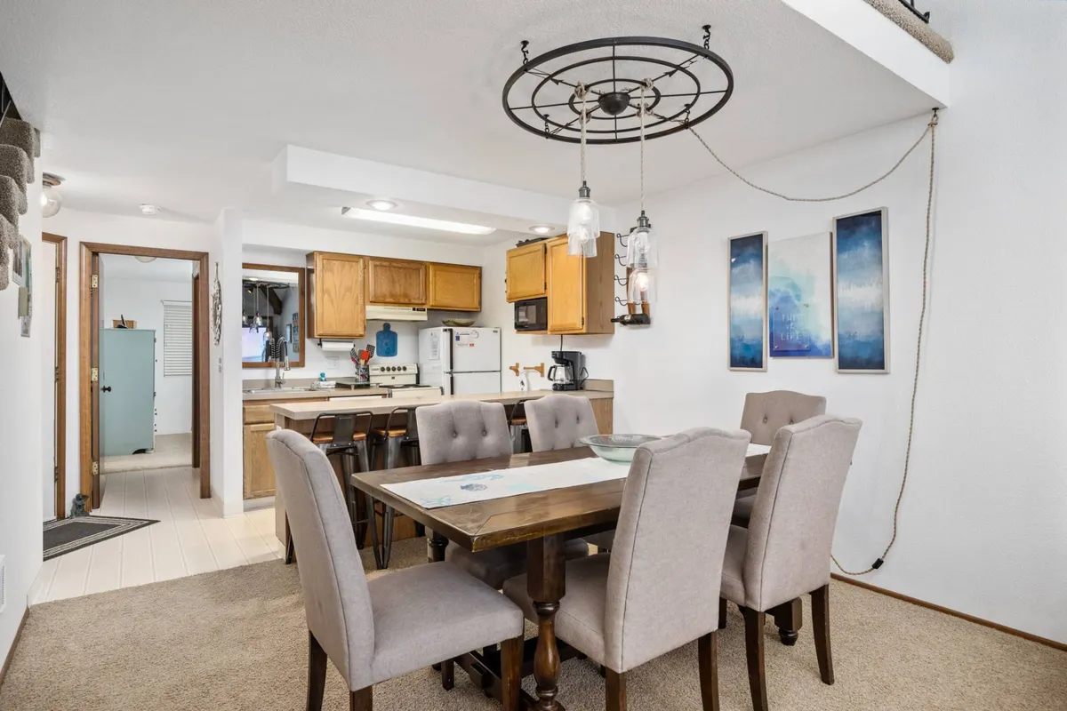 A warmly lit dining area featuring a six-person table positioned next to the kitchen area. The scene includes a stylish light fixture above the table, elegant wall art, and a kitchen bar visible in the background, inviting guests to gather and dine together.