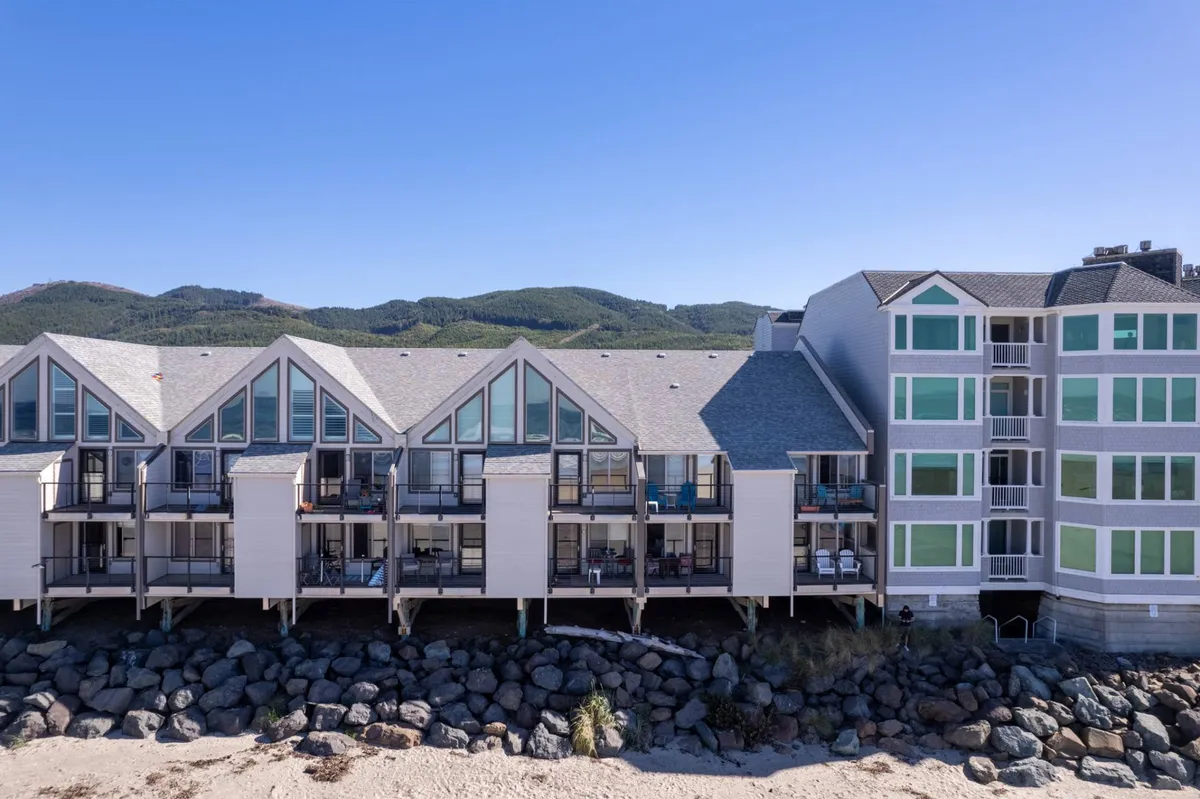 The exterior of a beachfront property with inviting balconies furnished for seating, set against a backdrop of the rocky shoreline and mountains. The balconies provide a private, front-row seat to breathtaking sunsets or peaceful mornings by the coast.