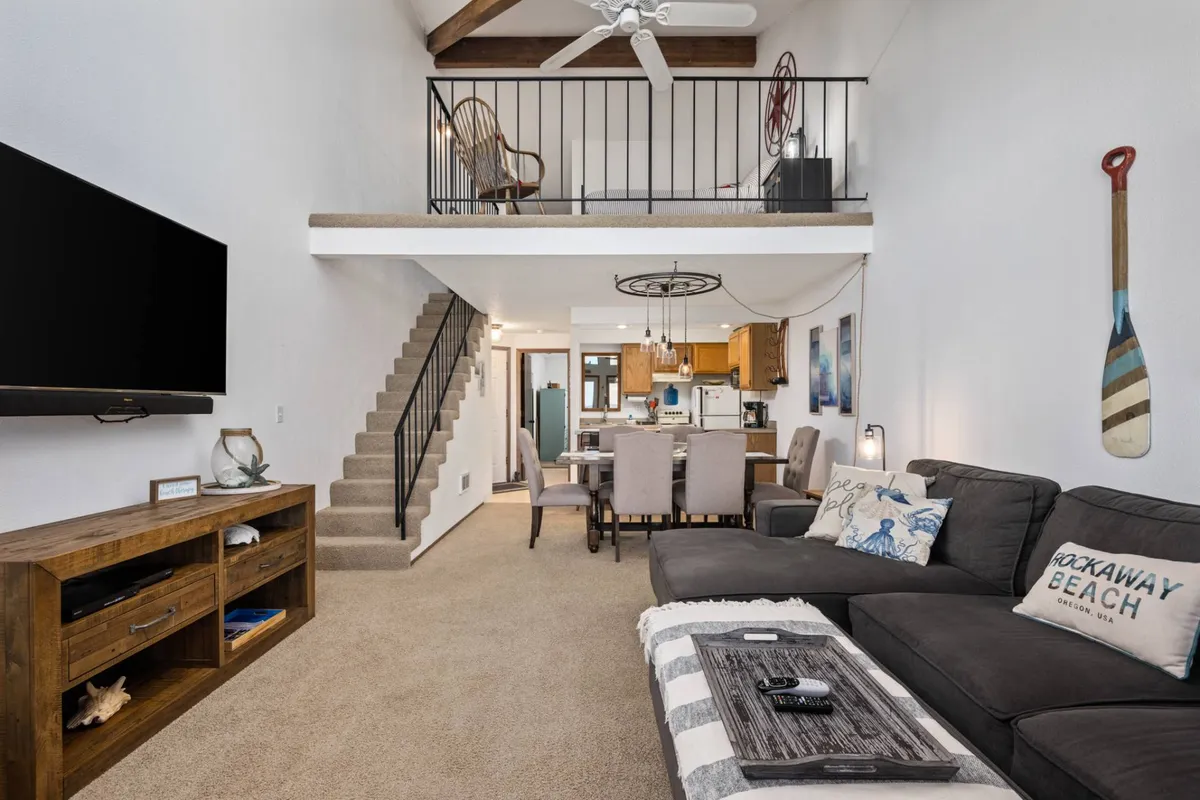 A welcoming open-concept living and dining space with a comfy sofa draped in a throw blanket, a large smart TV for movie nights, and a dining area adjacent to the staircase leading to a loft. The setting is casual yet refined, ideal for cozy evenings or entertaining guests.