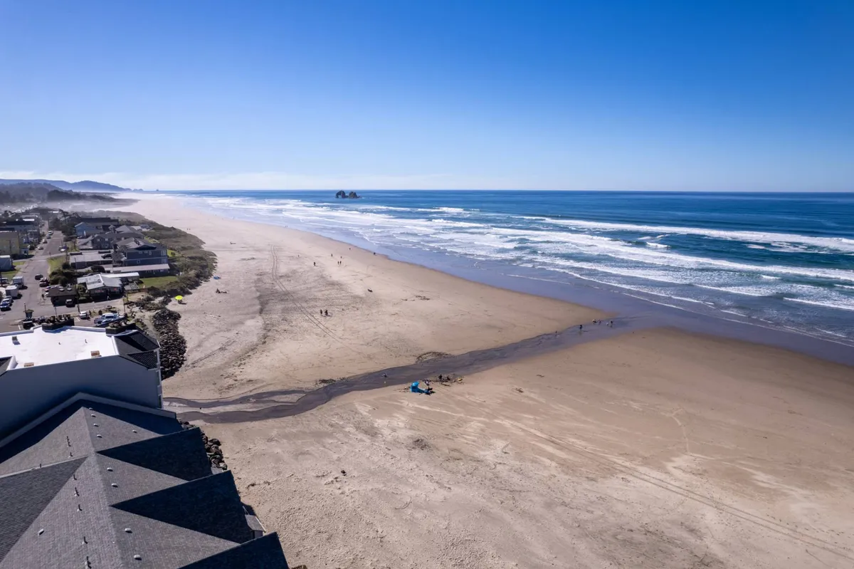 A wide-angle view of the expansive beach with gentle ocean waves, dotted with a few coastal homes nestled along the dunes. The scene is open and inviting, creating a sense of space and tranquility while also hinting at the proximity of accommodations to the shore.