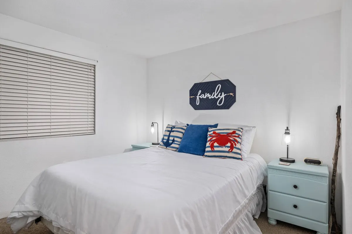 A cozy bedroom scene with blue and white linens, a decorative crab pillow, and a 'family' sign creates an inviting and restful environment. The decor is beach-themed, reinforcing the coastal location and the condo's comfortable, home-like ambiance.